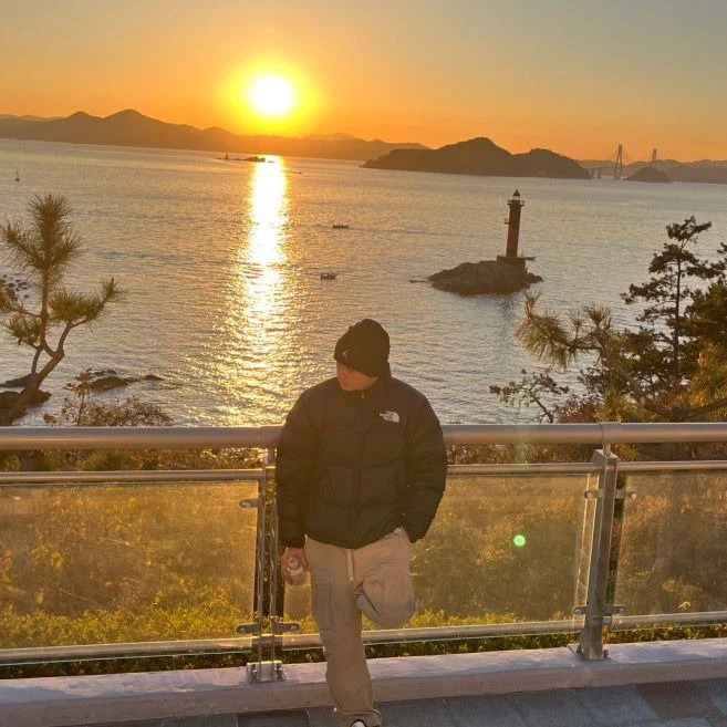 YANGLEE de pie junto a una baranda frente al mar al atardecer